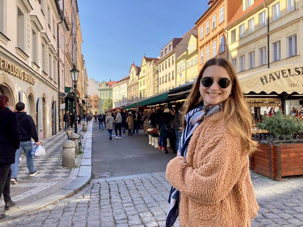 girl in a fluffy coat and sunglasses smiling at the camera in the old town of Prague with colorful buildings and a market in the background